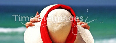 Girl sitting on sandy beach in the sun adjusting hat. Blue sky, blue sea red scarf. Spain.